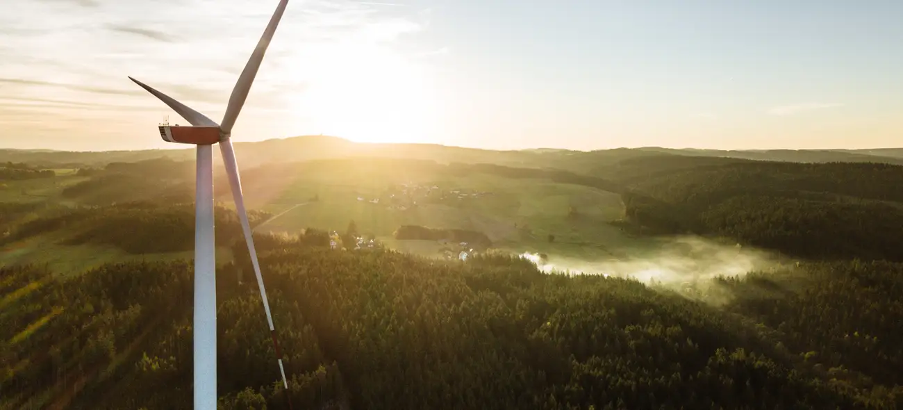 Nahaufnahme eines einzelnen Windrads bei Sonnenaufgang über bewaldetem Hügel (Querformat).
