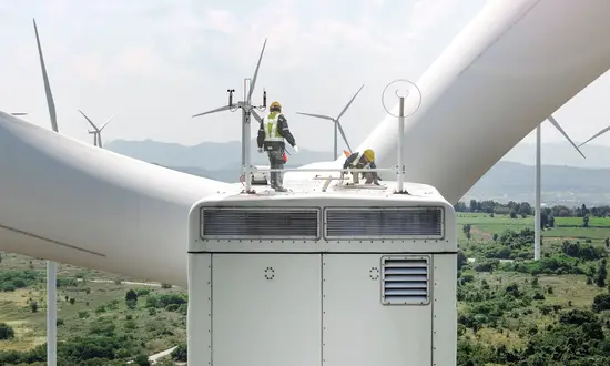 Techniker stehen auf einer Windkraftanlage und inspizieren einen weiteren Windpark in hügeliger Landschaft.